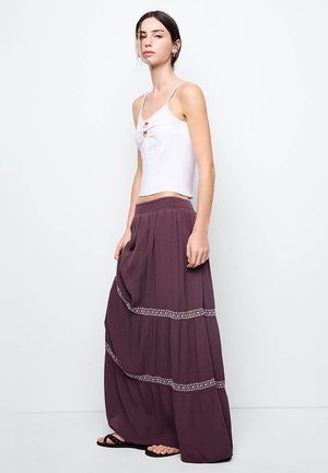 Jonge vrouw draagt een wit mouwloos topje en een lange paarse rok met patroonbanden, staand tegen een effen lichte achtergrond.