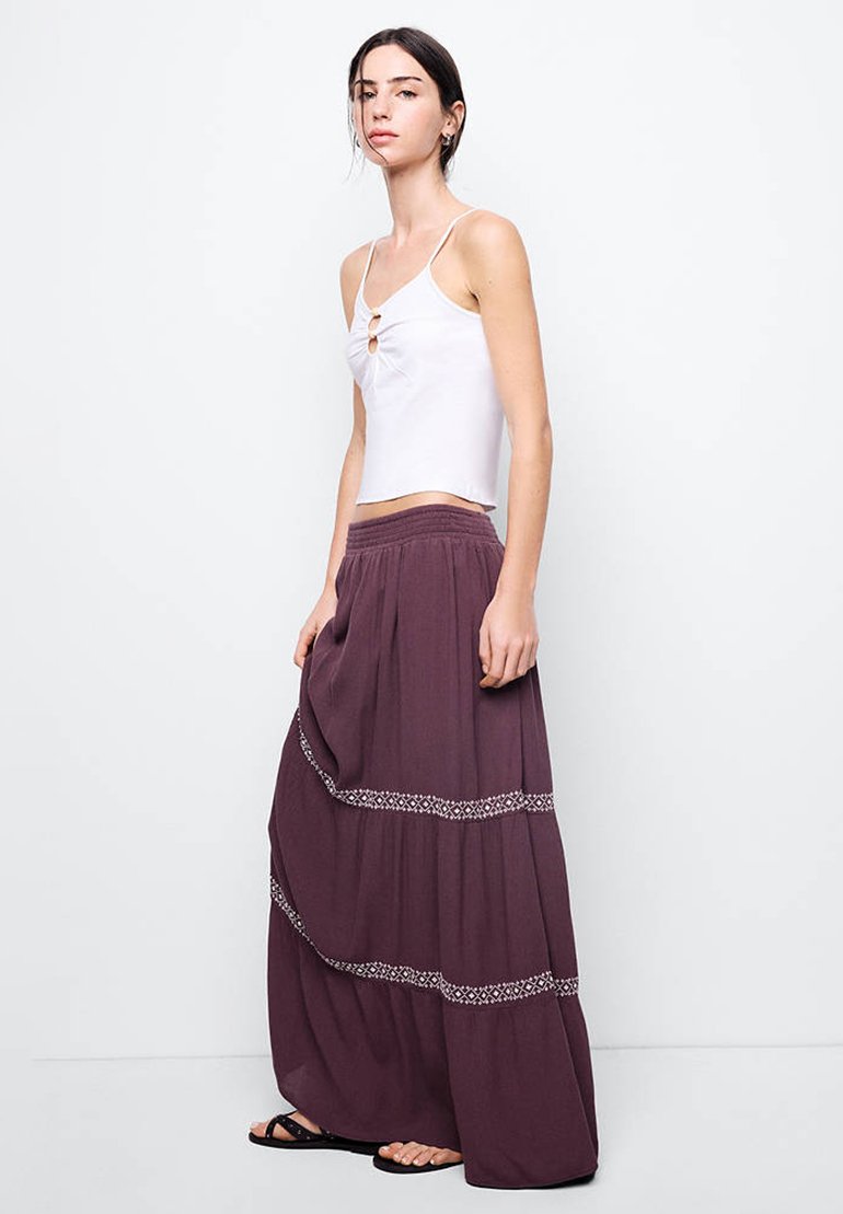 Jeune femme portant un haut blanc sans manches et une longue jupe violette avec des bandes à motifs, debout devant un fond clair uni.