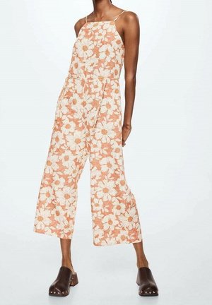 Kvinde iført en ærmeløs jumpsuit med vidde bukseben og orange og hvidt blomsterprint samt mørkebrune klodser med metalnitter.