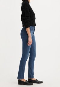 Suéter negro de punto trenzado, ajustado en la cintura; jeans azules de corte slim con costuras sutiles; mocasines negros con un acabado elegante.