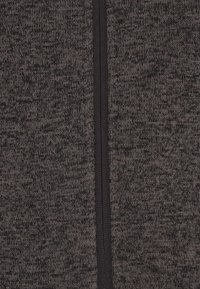 Sötétszürke kötött anyag strukturált mintával, központi fekete cipzárral. Az anyag puhanak és enyhén rugalmasnak tűnik.