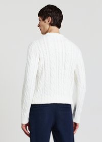 Weißer Strickpullover mit Zopfmuster, hohem Kragen und gerippten Bündchen, mit strukturierten Mustern. Kombiniert mit dunkelblauen Hosen.