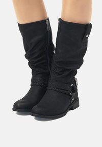 Botas negras hasta la rodilla, hechas de un tejido suave, con una correa trenzada, detalles en metal plateado y un tacón bajo cuadrado. Cierre con cremallera.