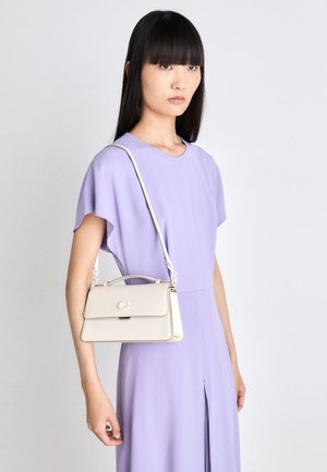 Mujer de cabello largo y oscuro con un vestido lavanda, llevando un pequeño bolso beige de bandolera sobre el hombro, sobre un fondo liso.