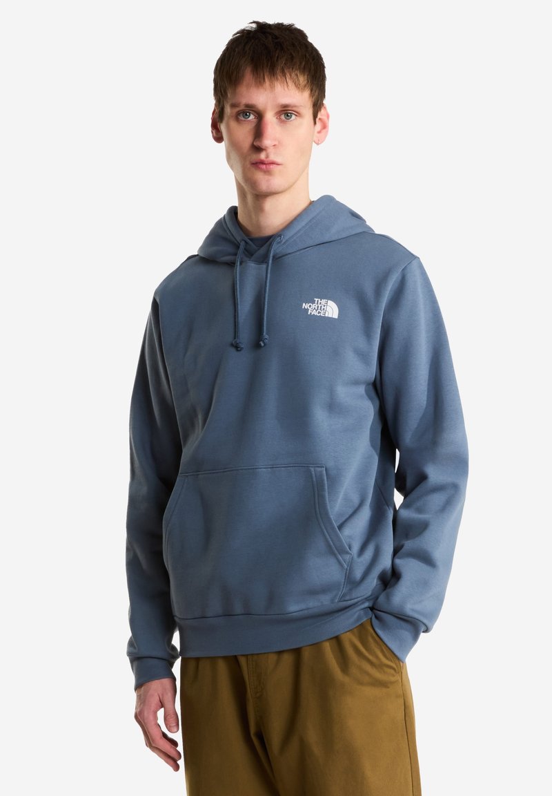 Jeune homme portant un sweat à capuche bleu The North Face avec une poche avant et un pantalon marron, debout devant un fond blanc.