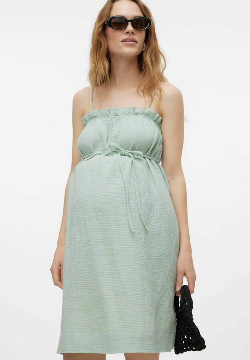 Jasnozielona sukienka ciążowa z fakturowanej tkaniny, z falbaną przy dekolcie i wiązaniem w pasie. Modelka trzyma czarną torebkę z koronkowym wzorem.