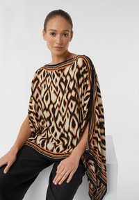 Wzorzysty poncho w kolorze pomarańczowym, czarnym i beżowym o płynnej formie. Posiada szeroką linię dekoltu oraz kontrastujące brzegi wzdłuż krawędzi.