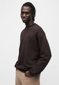 Sweat-shirt marron à col rond et poignets côtelés, avec un petit logo brodé sur la poitrine, associé à un pantalon rayé beige.