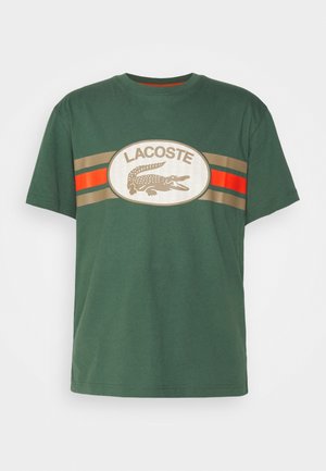 Zielona bawełniana koszulka z logo w kształcie owalnym na środku, z napisem "LACOSTE", grafiką krokodyla oraz poziomymi paskami w beżu i czerwieni.