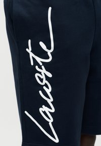 Σκούρα μπλε σορτς με λευκό καλλιγραφικό λογότυπο "Lacoste" εκτυπωμένο κάθετα κατά μήκος του αριστερού μηρού.