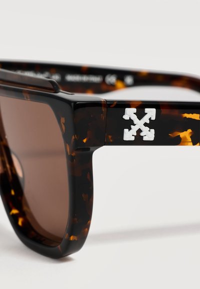 Gros plan sur des lunettes de soleil écaille de tortue marron avec des verres rectangulaires et un logo croix blanche à quatre flèches sur la branche.