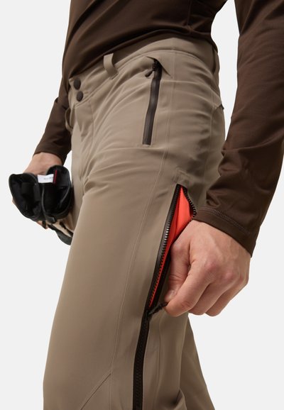 Béžové outdoorové kalhoty s bočními zipovými kapsami a červenou podšívkou; nošené s hnědým dlouhým rukávem. Ruka držící kalhoty poblíž zipu.