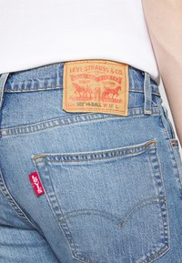 Vista trasera de unos jeans azules de Levi's con parche de cuero marrón visible y etiqueta roja de Levi's en el bolsillo, combinados con una camisa blanca.