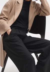 Manteau en mélange de laine beige sur un haut noir, associé à un pantalon noir taille haute. Textures lisses, avec une coupe décontractée et des plis subtils.