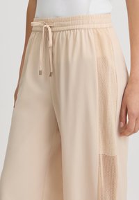 Pantaloni sportivi beige con vita regolabile, dotati di un pannello laterale in rete testurizzata. Realizzati in materiale leggero e liscio.
