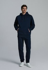 Granatowy dres z miękkiej tkaniny, składający się z bluzy z kapturem oraz spodni. Bluza ma przednią kieszeń i elastyczne mankiety. Zestaw połączony z białymi trampkami.