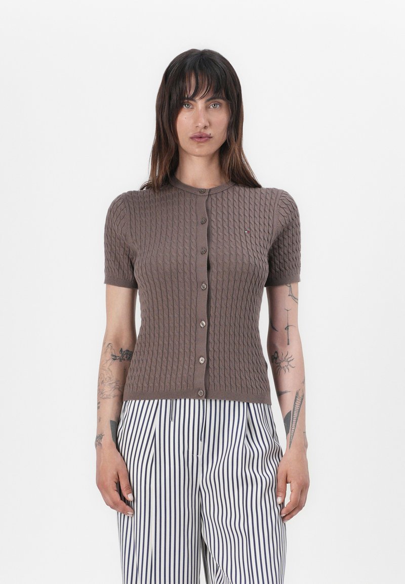 Cardigan marrone a coste con maniche corte, scollatura rotonda e chiusura con bottoni. Presenta un piccolo logo ed è abbinato a pantaloni a righe.
