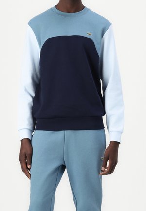 Homme portant un sweat-shirt bleu à blocs de couleur avec des manches blanches et un pantalon de jogging bleu assorti, les deux avec de petits logos de marque.