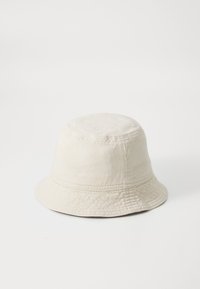 Chapeau seau en coton beige avec un dessus arrondi et un large bord tombant. Détails de couture le long de la base et de la couronne.
