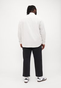 Wit langrimpelig shirt met lichte verticale strepen, gecombineerd met donkere broeken en witte sneakers. Eenvoudig ontwerp, met zichtbare geknoopte manchetten.