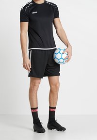 Černý sportovní dres s šedými akcenty a logem, doplněný černými šortkami, černými kopačkami a ponožkami se červenými pruhy. Drží modrobílý fotbalový míč.