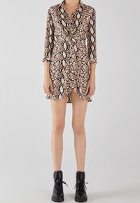 Abito camicia con stampa serpe, colletto, maniche lunghe e vita annodata. Il tessuto presenta tonalità di beige, marrone e nero. Indossato con stivaletti neri alla caviglia.