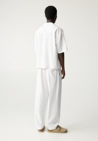 Wit overhemd met korte mouwen en losse broek gemaakt van lichtgewicht stof. Beige instapschoenen met bandje, gezien van achteren.