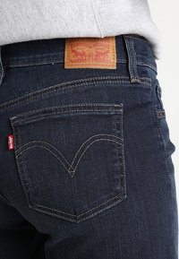 Donkerblauwe spijkerbroek met een leren merkpatch aan de achterkant, zichtbare stiksels en een hartvormig zakdetail aan de rechterkant.