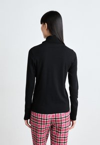 Schwarzer Strickpullover mit hohem Kragen, langen Ärmeln und gerippten Bündchen, kombiniert mit rosa Hahnentrittmuster-Hosen.