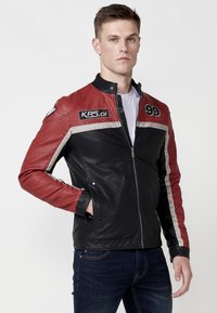 Čierna a červená kožená motocyklistická bunda so striebornými pruhmi, postaveným golierom, zipsom a vyšitými nášivkami vpredu.