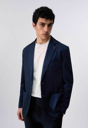 Giovane uomo con capelli neri ricci che indossa un blazer blu navy sopra una camicia bianca, in piedi contro uno sfondo grigio chiaro uniforme.