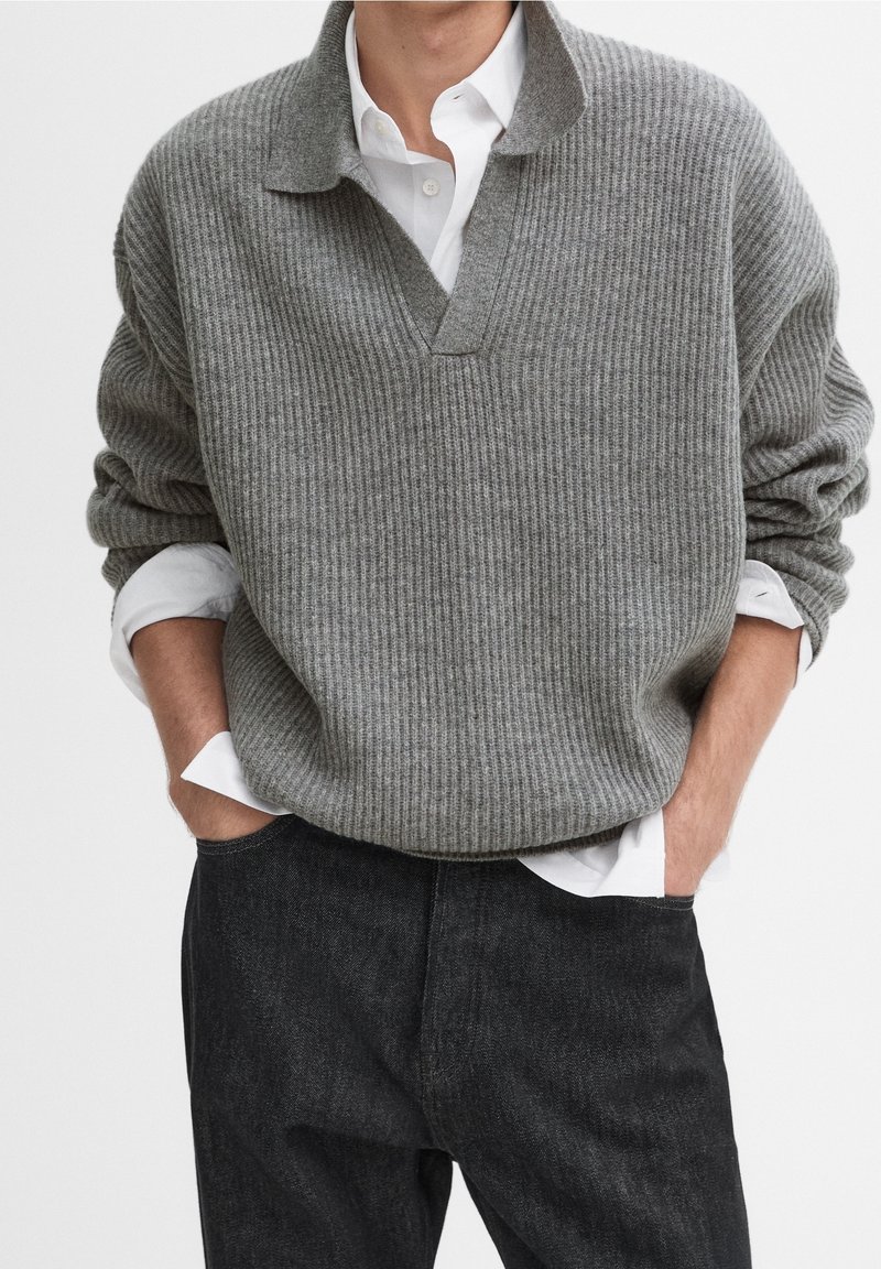 Homme portant un pull gris côtelé par-dessus une chemise blanche aux manches retroussées, les mains dans les poches d'un pantalon noir.