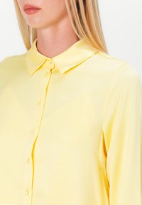 Yellow satin blouse featuring a classic collar, button closure, and long sleeves; smooth texture with a soft sheen.