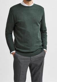 Ciemnozielony sweter dzianinowy z okrągłym dekoltem, ściągaczami na rękawach i u dołu, w połączeniu z szarymi spodniami o teksturowanej fakturze. Brak wyraźnych wzorów lub akcentów.