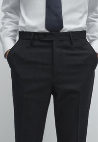 Dunkelgraue, taillierte Hose mit strukturiertem Finish, Knopfverschluss und Seitentaschen, kombiniert mit einem weißen gestreiften Hemd und einer dunklen Krawatte.