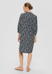 Schwarz-weißes Kleid mit geometrischem Muster, langen Ärmeln, geradem Schnitt und seitlichen Schlitzungen. Getragen mit flachen Sandalen.