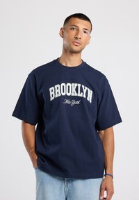 T-paita tummansinisestä puuvillasta, jossa on valkoisilla kirjaimilla "BROOKLYN New York", lyhyet hihat ja rento istuvuus.