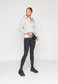 Νεαρή γυναίκα με γκρι φούτερ με κουκούλα και μαύρο κολάν, κρατώντας μαύρο παγούρι, στέκεται μπροστά σε φωτεινό απλό φόντο, κοιτάζοντας προς το πλάι.