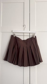 Brauner Faltenrock mit einem Knopfdetail, hängt an einem Kleiderbügel vor einem weißen Hintergrund. Verfügt über klare Linien und eine taillierte Passform.