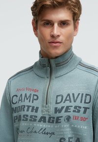 Hellblauer Pullover mit Reißverschluss am Kragen und strukturierter Strickoptik, mit grafischen Drucken in Dunkel- und Rottönen. Akzente durch gerippten Kragen und Schulterstreifen.