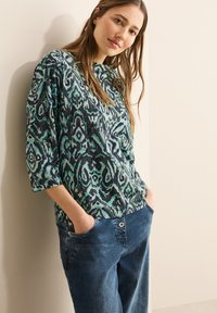 Blouse multicolore avec un motif abstrait en vert et bleu, présentant des manches trois-quarts et une coupe décontractée, associée à un pantalon en denim bleu.