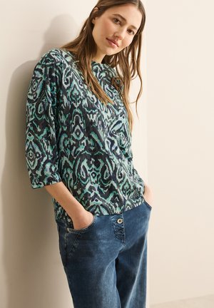 Blouse multicolore avec un motif abstrait en vert et bleu, présentant des manches trois-quarts et une coupe décontractée, associée à un pantalon en denim bleu.
