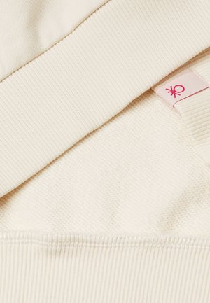 Tessuto color crema con superficie testurizzata, dotato di scollatura a coste e piccolo logo etichetta rosa sul lato.
