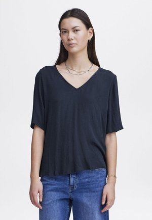 Zwarte V-hals top met losse korte mouwen, gemaakt van een gladde stof. Gecombineerd met blauwe denim jeans, gelaagd met zilveren kettingen.