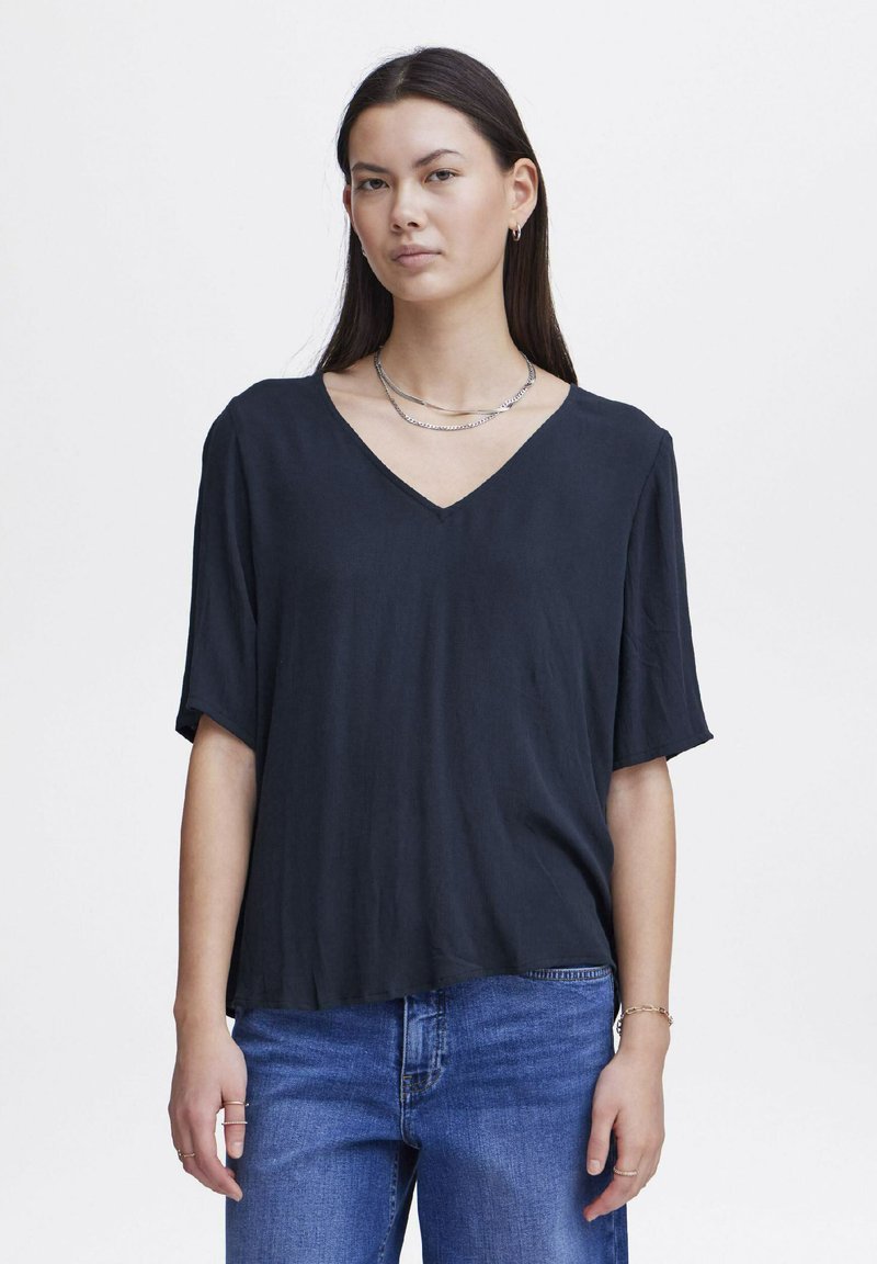 Haut noir à col en V avec des manches courtes amples, fabriqué en tissu lisse. Assorti à un jean en denim bleu, superposé avec des colliers en argent.