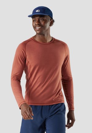 Junger Mann mit rostfarbenem Langarmhemd, marineblauer Hose und marineblauer Mütze, der lächelnd vor einem schlichten grauen Hintergrund geht.