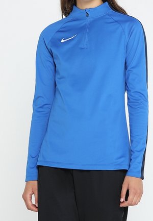 Osoba ubrana w niebieską sportową bluzę Nike z długim rękawem i kołnierzem na suwak oraz czarne spodnie, stoi na tle jasnego, jednolitego tła.