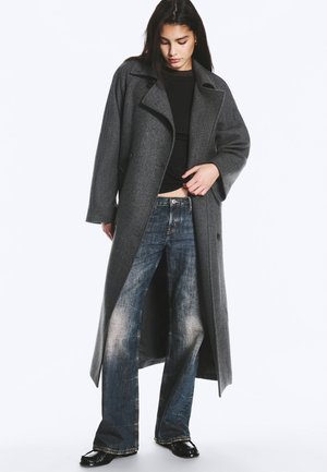 Une femme présente un long manteau gris anthracite porté sur un haut noir, un jean délavé à jambes larges et des chaussures noires, sur un fond blanc uni.