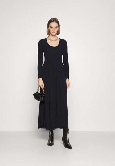Robe noire à manches longues avec un col rond, corsage ajusté et jupe fluide. Portée avec des bottines et un petit sac à main noir.