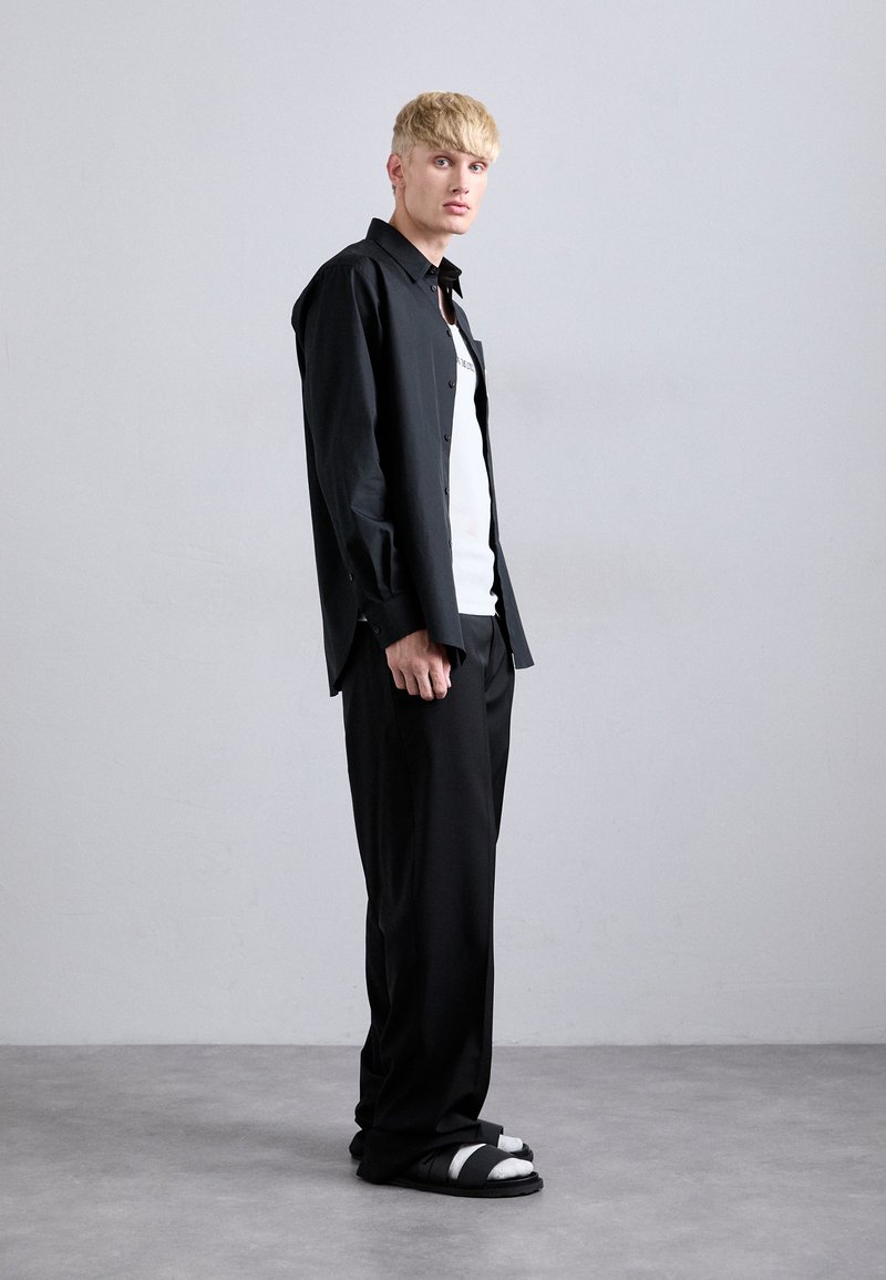 Young man standing against a plain wall wearing black wide-leg pants, black open shirt over a white tank top, and black sandals with white socks.